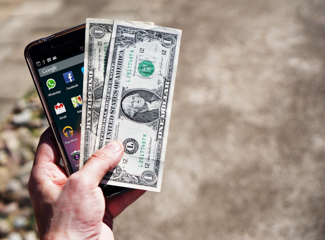 Close-up of a hand holding US dollar bills and a smartphone outdoors, showcasing financial technology.